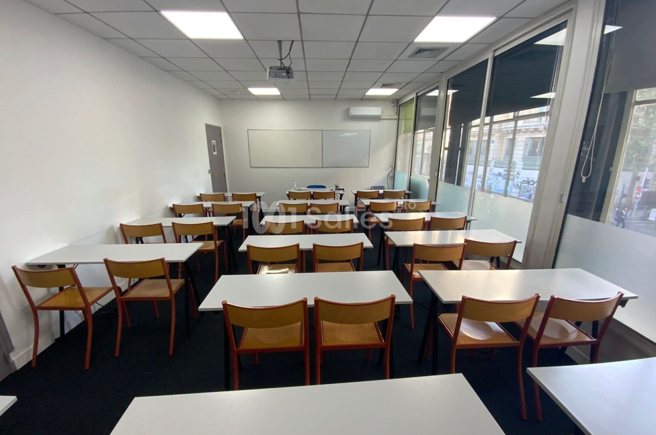 Salle de classe vide avec des tables blanches, des chaises en bois, un tableau blanc et des fenêtres donnant sur l'extérieur.