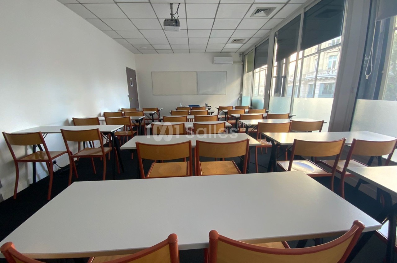 Salle de classe vide avec des tables blanches, des chaises en bois et un tableau blanc au fond, éclairée par des fenêtres.