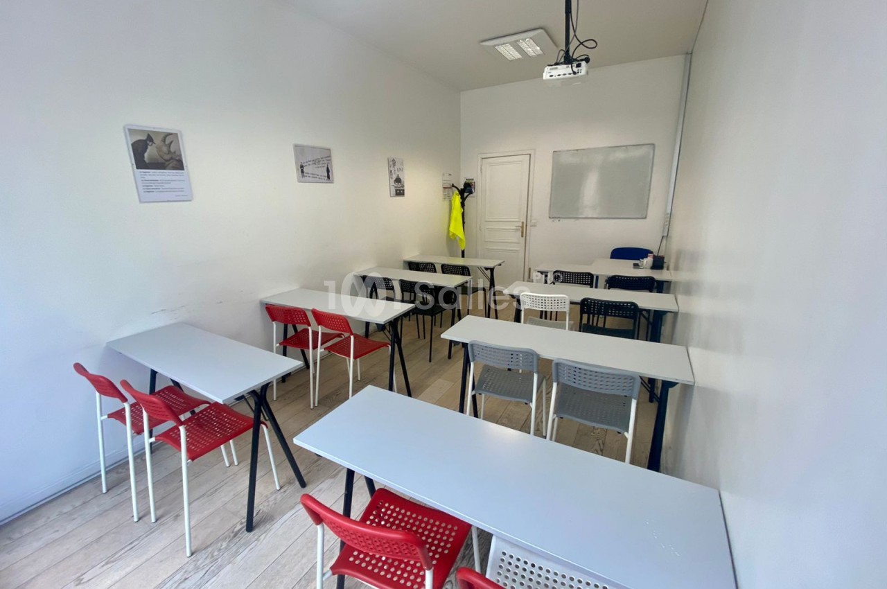 Salle de formation avec des tables blanches, des chaises rouges et noires, un tableau blanc et un projecteur au plafond.