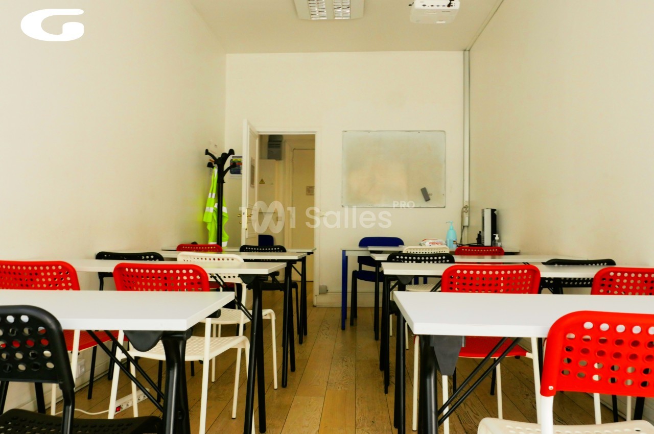 Salle de classe lumineuse avec des tables blanches, chaises rouges et noires, tableau blanc et projecteur au plafond.