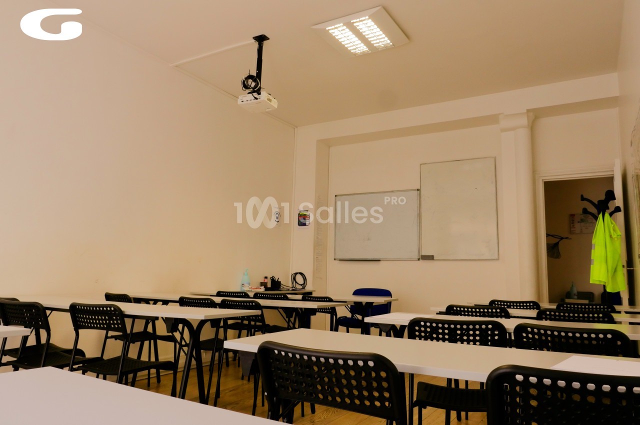 Salle de classe vide avec des tables, des chaises noires, un tableau blanc et un projecteur au plafond.