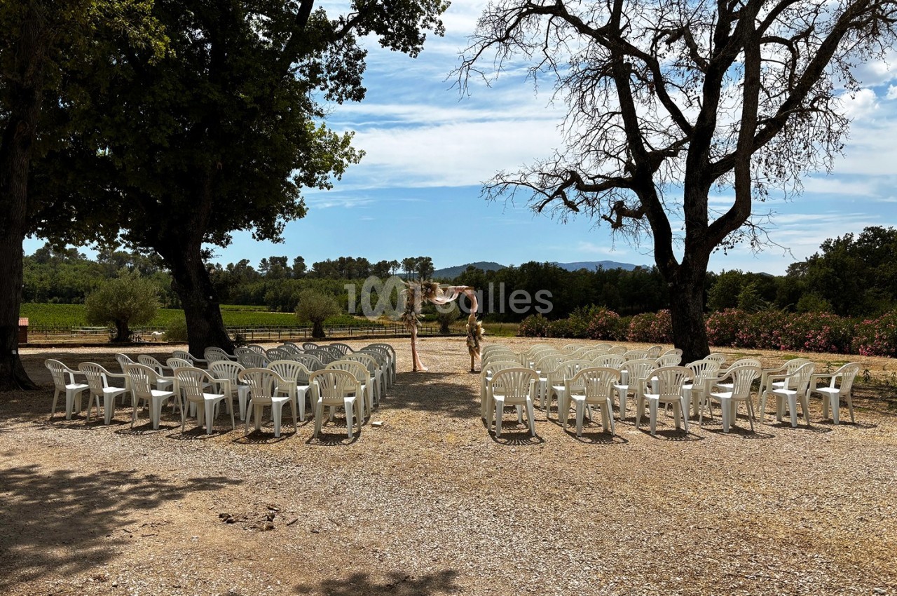 Chaises blanches disposées en rangées en plein air, face à une arche décorée, entourées d'arbres et de végétation.
