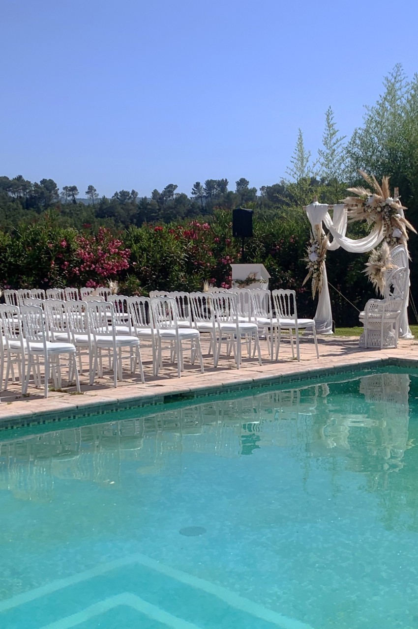 Chaises blanches alignées près d'une piscine, face à une arche décorée pour une cérémonie en extérieur.