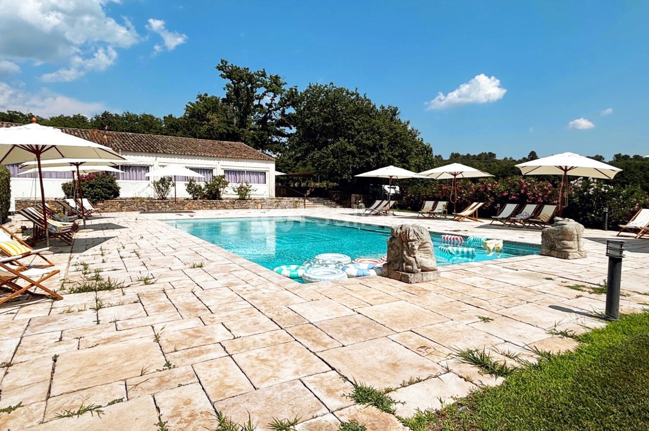 Piscine extérieure entourée de chaises longues, parasols et végétation, sous un ciel bleu dégagé.