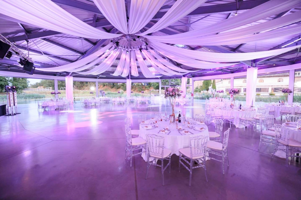 Salle de réception spacieuse avec tables rondes dressées, chaises transparentes et éclairage violet sous un plafond drapé.
