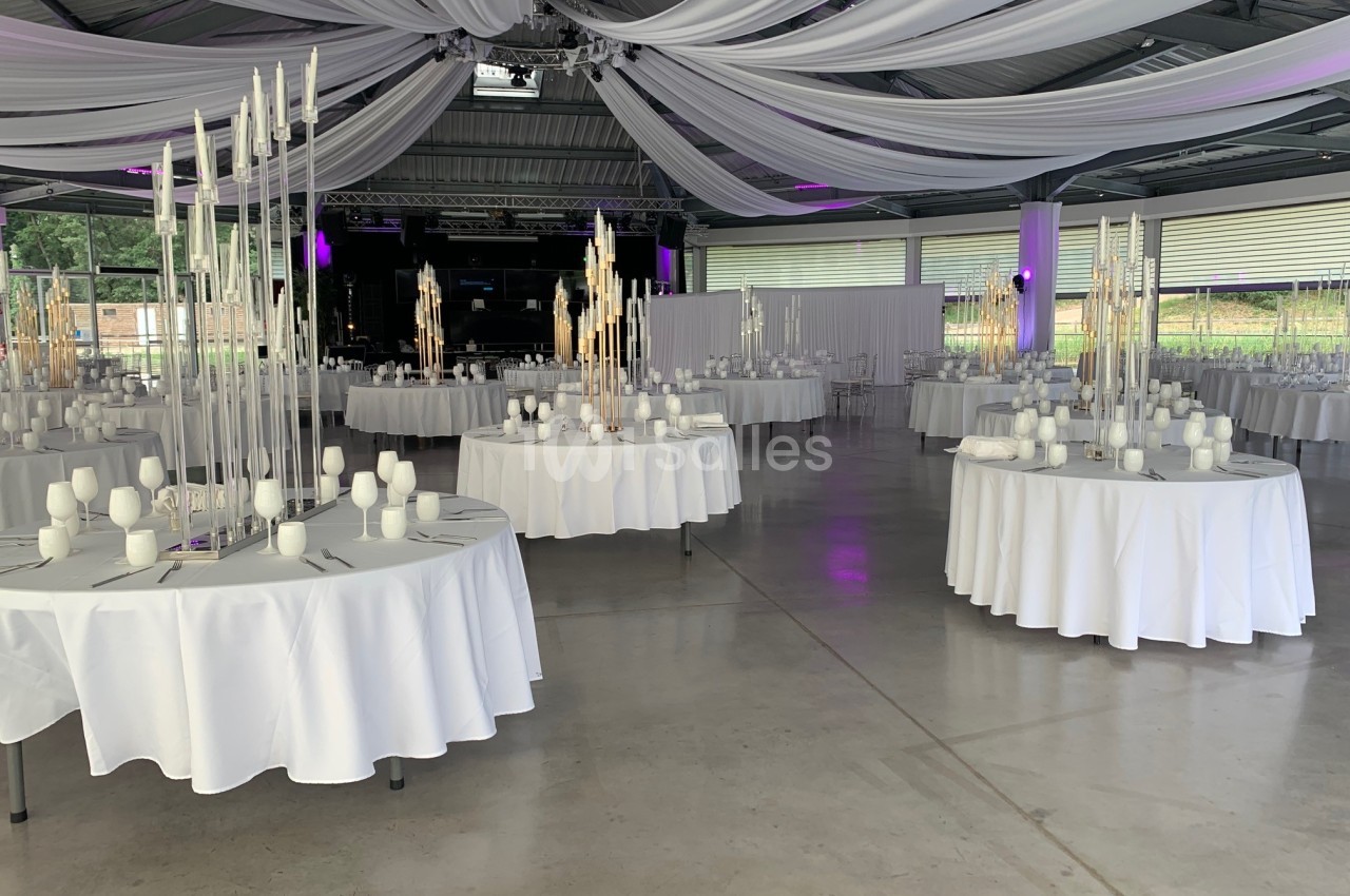 Salle de réception décorée avec des tables rondes, nappes blanches, chandeliers et drapés suspendus au plafond.