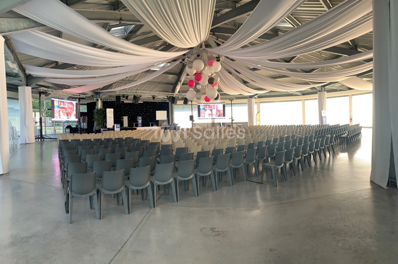 Salle de conférence décorée de voilages blancs, avec des rangées de chaises grises et une scène équipée d'écrans.