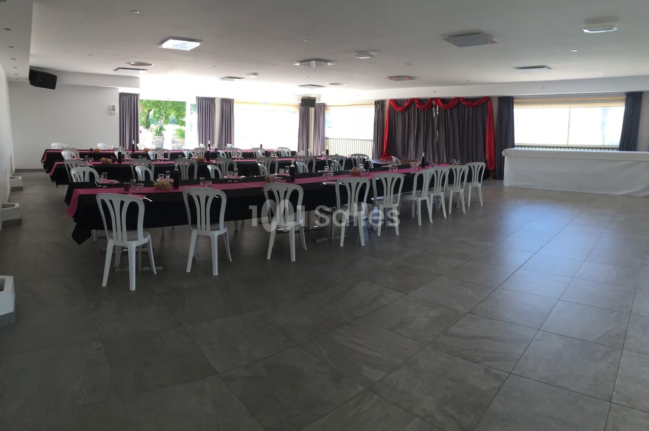 Salle de réception lumineuse avec tables dressées en noir et rose, chaises blanches et rideaux violets.