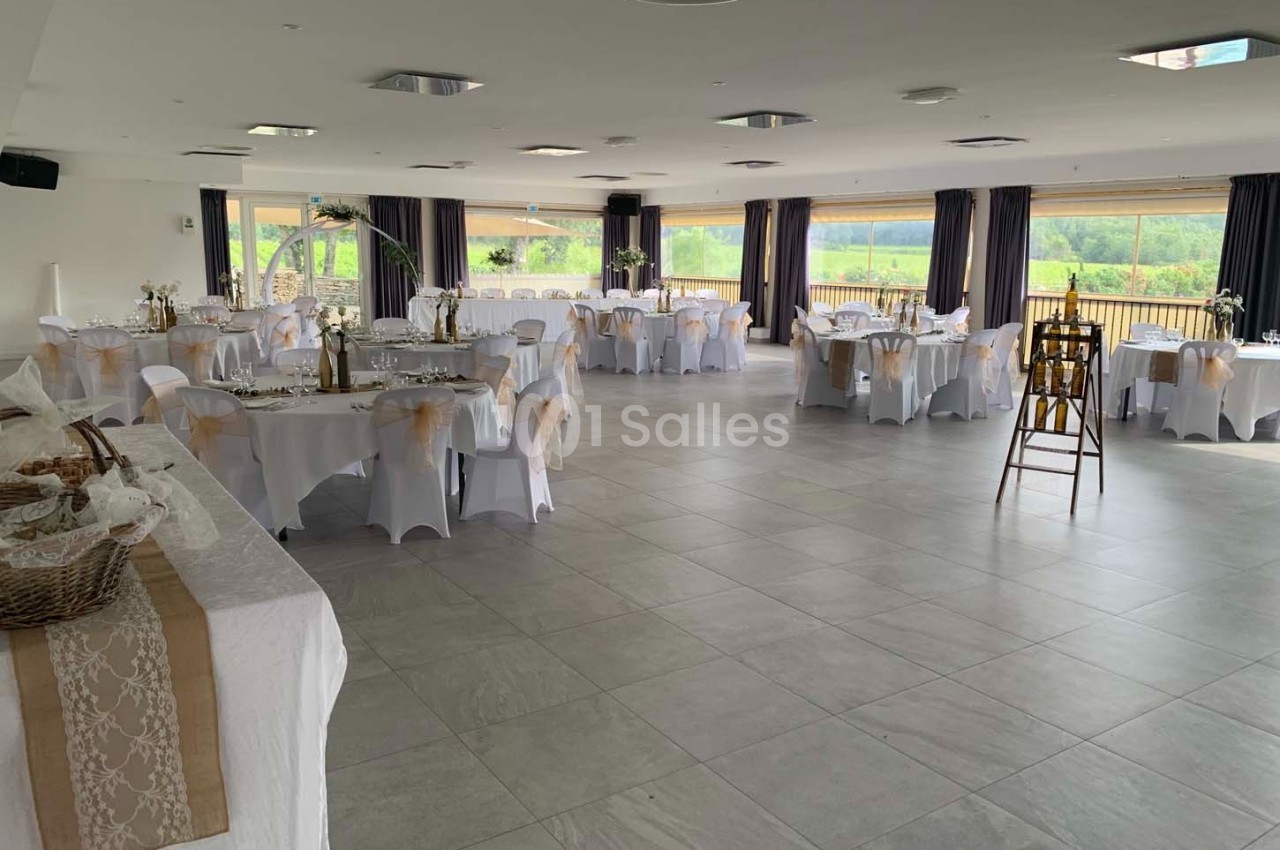 Salle de réception lumineuse avec tables rondes décorées, nappes blanches et grandes baies vitrées donnant sur un paysage…