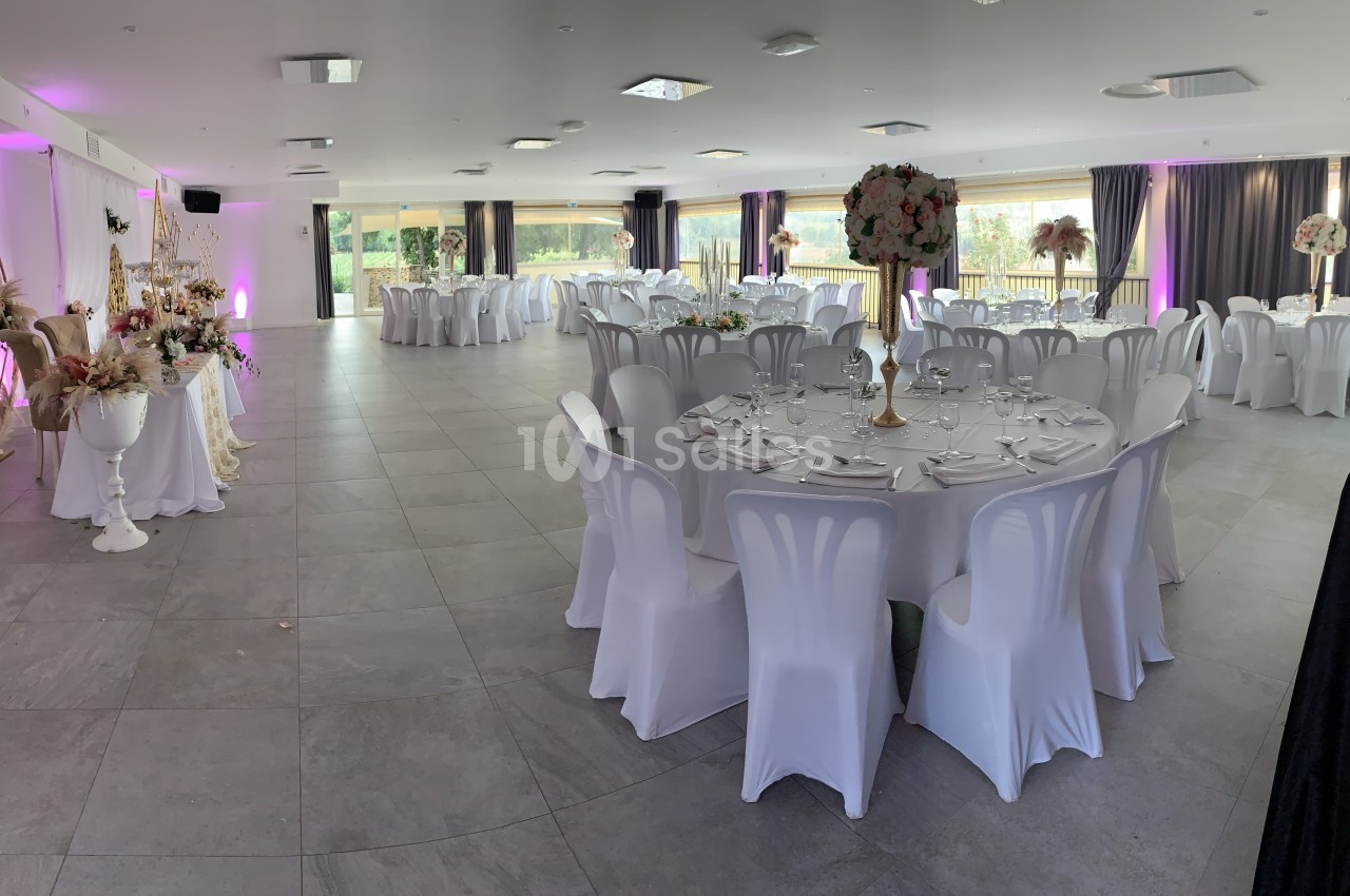 Salle de réception décorée avec des tables rondes dressées, des chaises blanches et des arrangements floraux élégants.