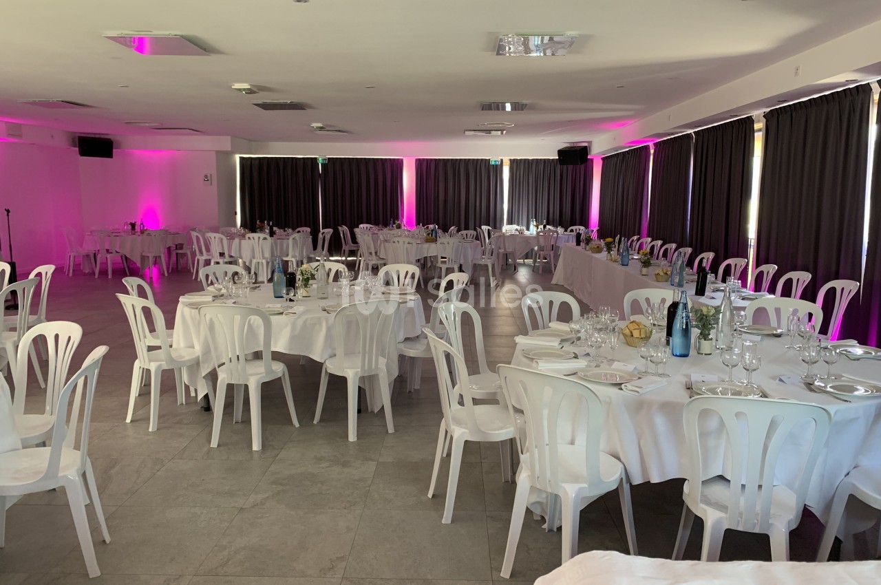 Salle de réception avec tables rondes et rectangulaires dressées, éclairage tamisé et rideaux sombres.