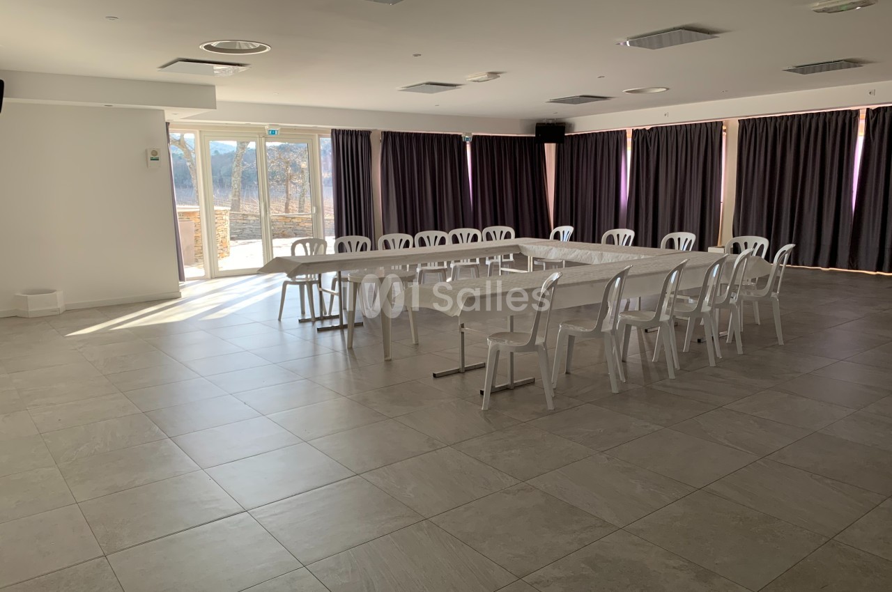Salle lumineuse avec des tables disposées en U, entourées de chaises blanches, et des rideaux fermés sur les fenêtres.