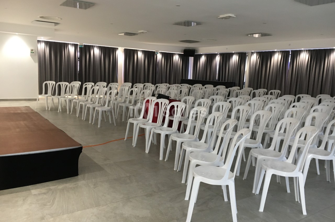 Salle de conférence vide avec des rangées de chaises blanches en plastique et une petite scène à gauche.