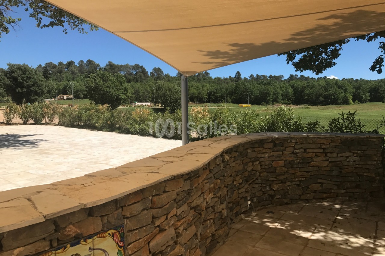 Mur en pierre courbé sous une toile d'ombrage, avec vue sur un espace vert et des arbres en arrière-plan.