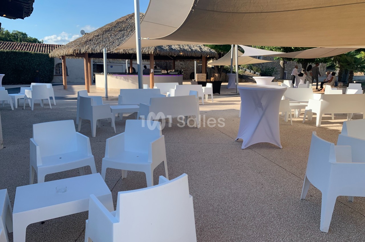 Espace extérieur avec mobilier blanc, tables et chaises sous des voiles d'ombrage près d'un bar au toit de chaume.