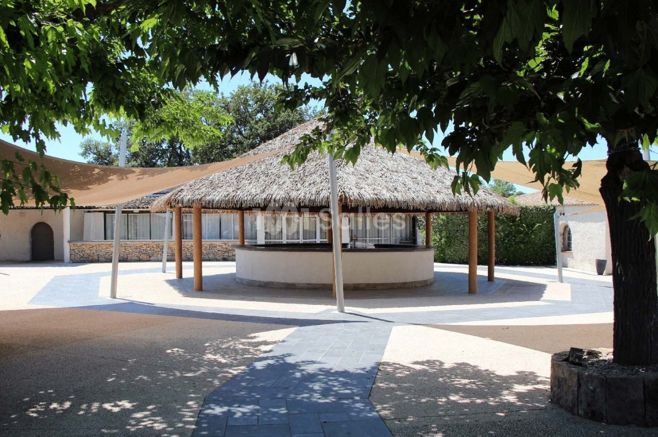 Kiosque en toit de chaume entouré d'arbres, situé dans une cour pavée avec des voiles d'ombrage.