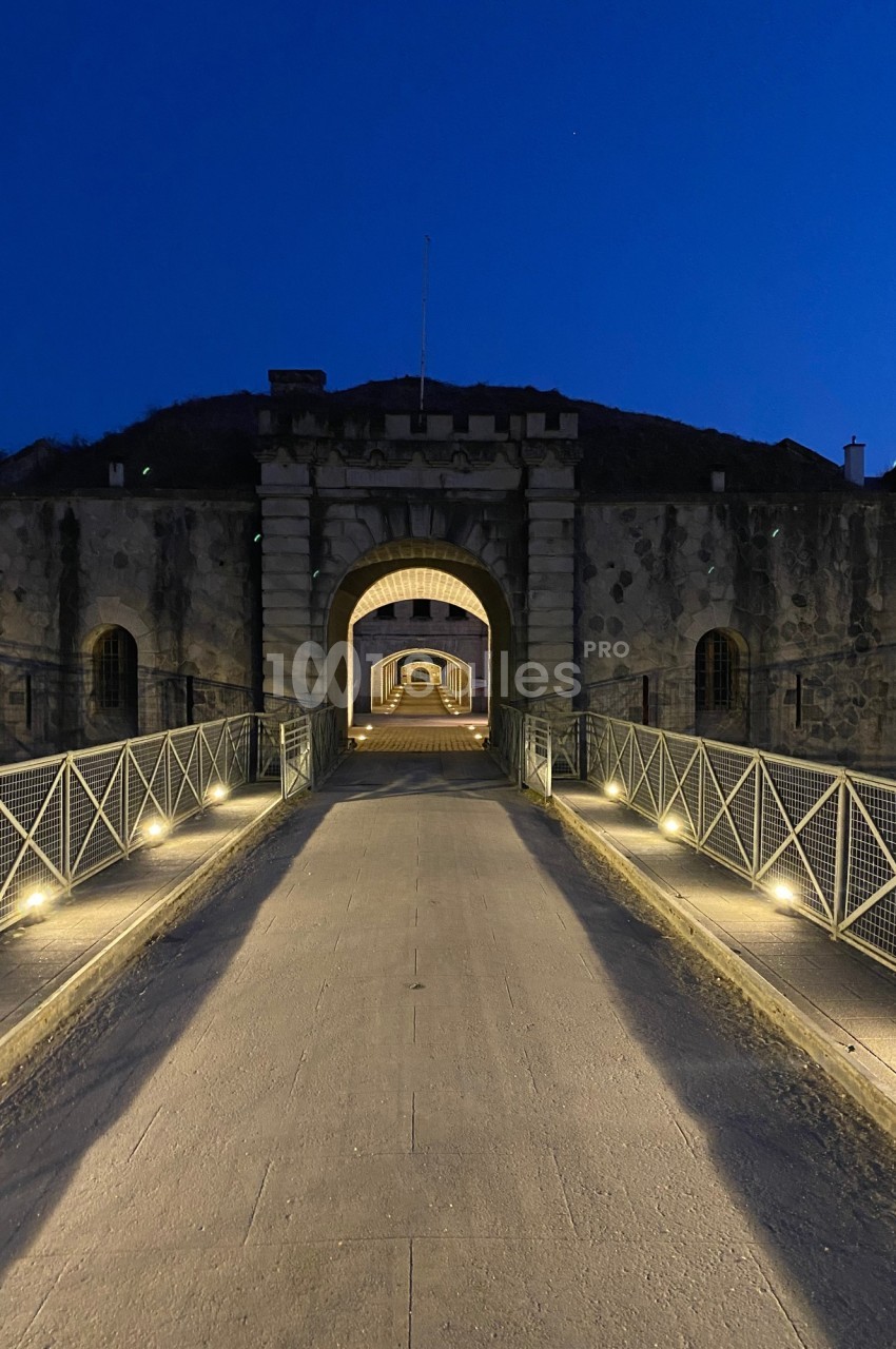 Passerelle éclairée menant à une entrée voûtée d'un fort en pierre, photographiée à la tombée de la nuit.