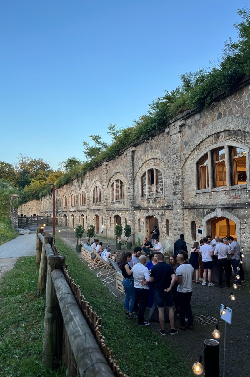 Groupe de personnes rassemblées devant un bâtiment en pierre avec des arches, dans un cadre verdoyant en soirée.