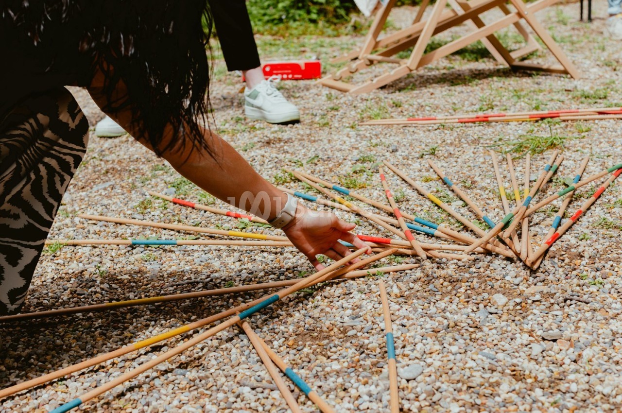 Personne ramassant des baguettes de mikado en bois coloré éparpillées sur un sol de gravier.