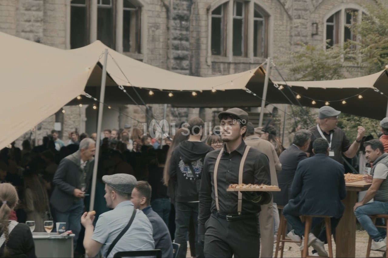 Serveur portant un plateau dans un événement en plein air sous des tentes, avec des invités assis et debout.