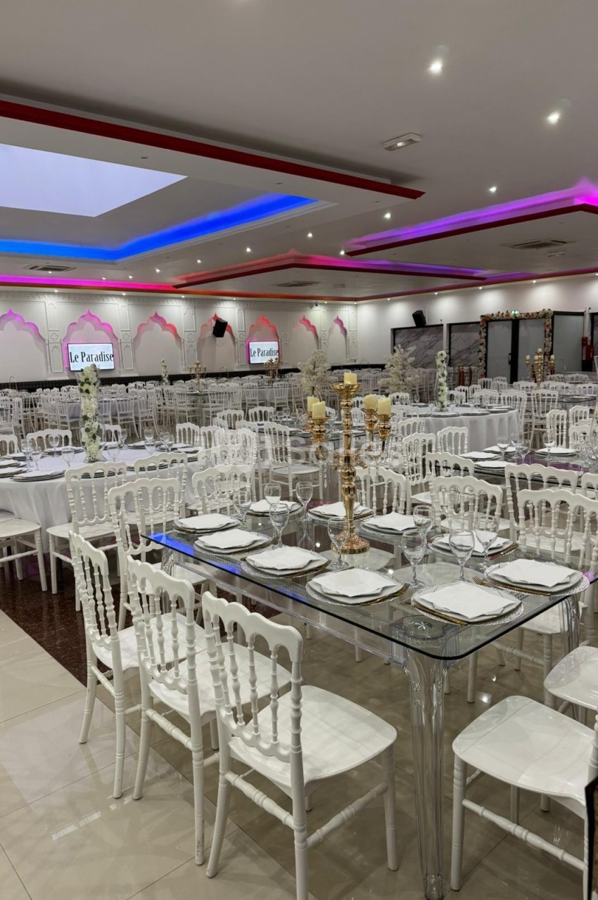 Salle de réception décorée avec des tables dressées, chaises blanches et éclairage coloré au plafond.
