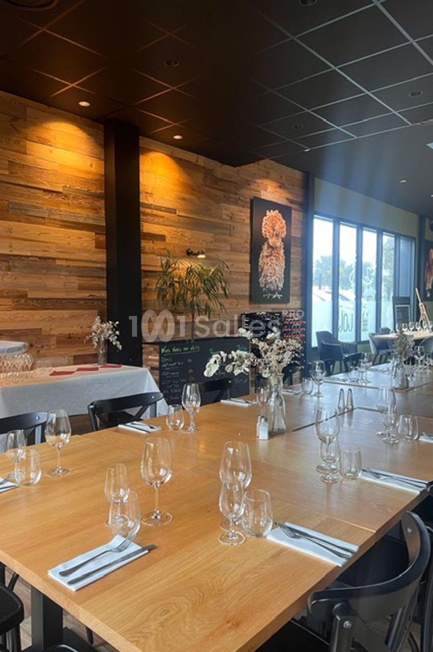 Salle de restaurant moderne avec tables en bois dressées, décor mural en bois et grandes baies vitrées.
