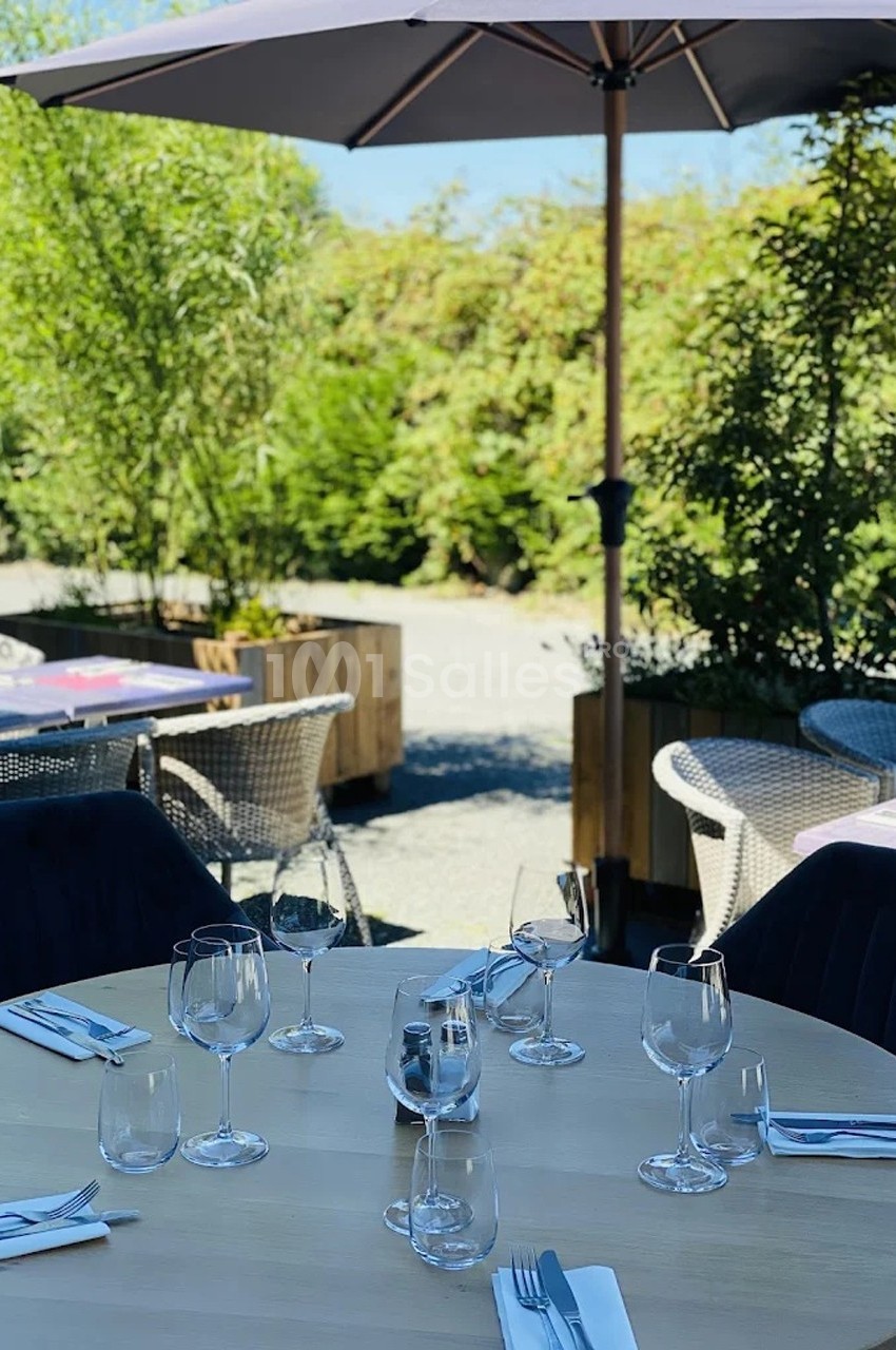 Table dressée en extérieur avec verres et couverts, entourée de chaises, sous un parasol et près de végétation.