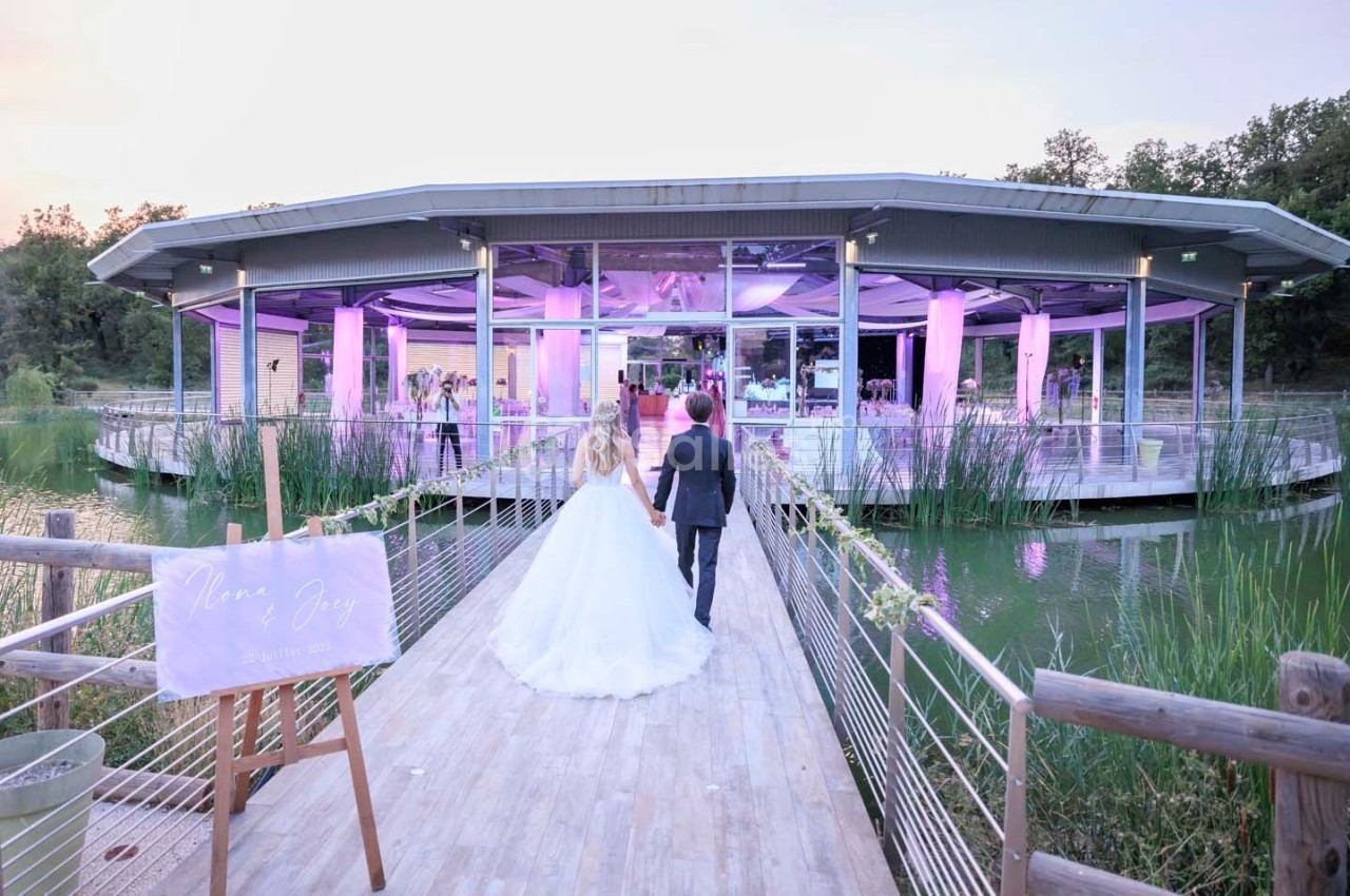 Un couple marche main dans la main sur une passerelle menant à une salle de réception illuminée en rose, entourée d'eau.