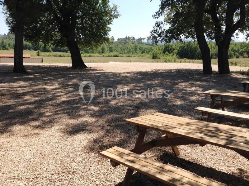 Tables de pique-nique en bois sous l'ombre d'arbres, avec une vue dégagée sur un champ et des arbres au loin.