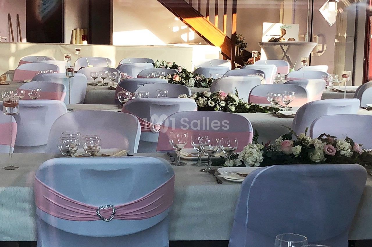 Salle de réception décorée avec des chaises blanches ornées de nœuds roses et des tables dressées avec des fleurs.