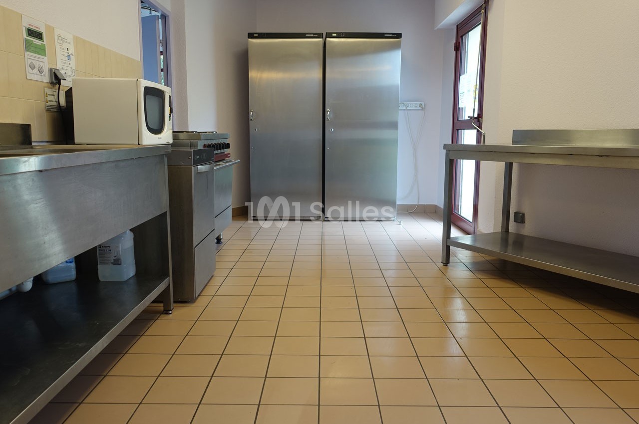 Cuisine professionnelle avec sol carrelé, plans de travail en inox, micro-ondes et deux grands réfrigérateurs en arrière…