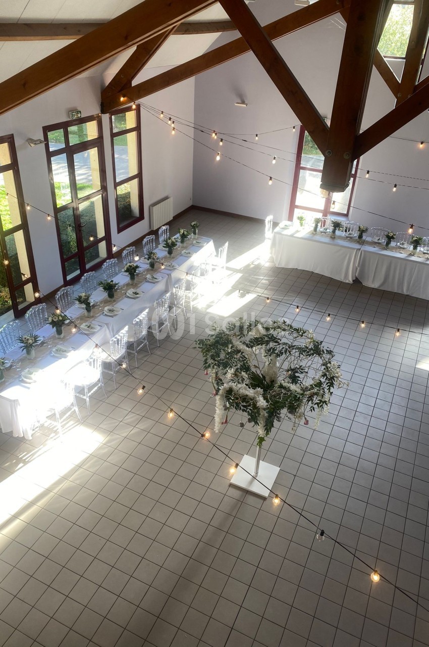 Salle lumineuse décorée pour un événement, avec tables alignées, chaises transparentes et guirlandes suspendues.