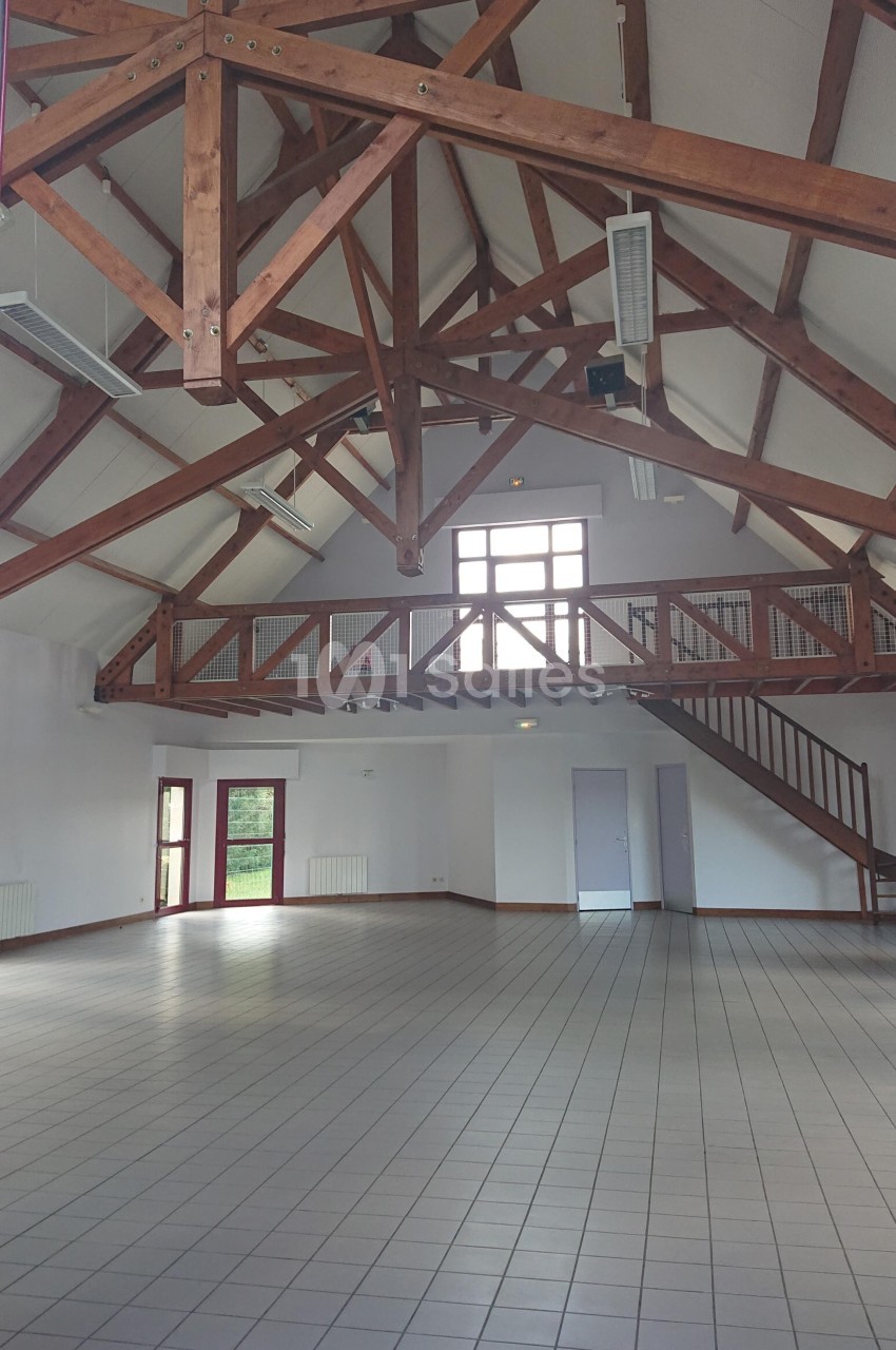 Salle spacieuse avec charpente en bois apparente, carrelage au sol, mezzanine et grandes fenêtres laissant entrer la lumière.