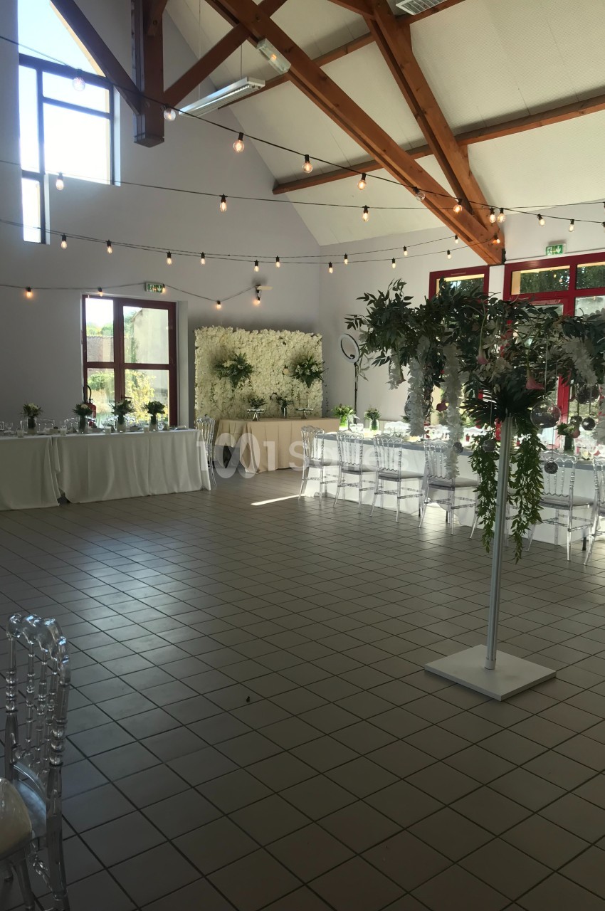 Salle de réception décorée pour un événement, avec tables dressées, chaises transparentes et guirlandes lumineuses…