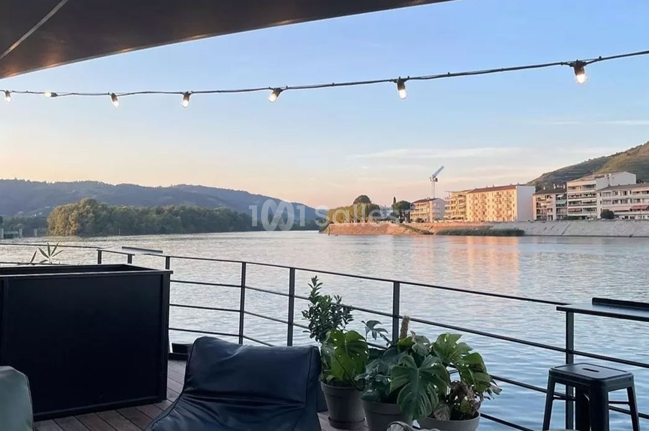 Vue depuis une terrasse en bois avec des plantes, donnant sur une rivière bordée de bâtiments et de collines au coucher du…
