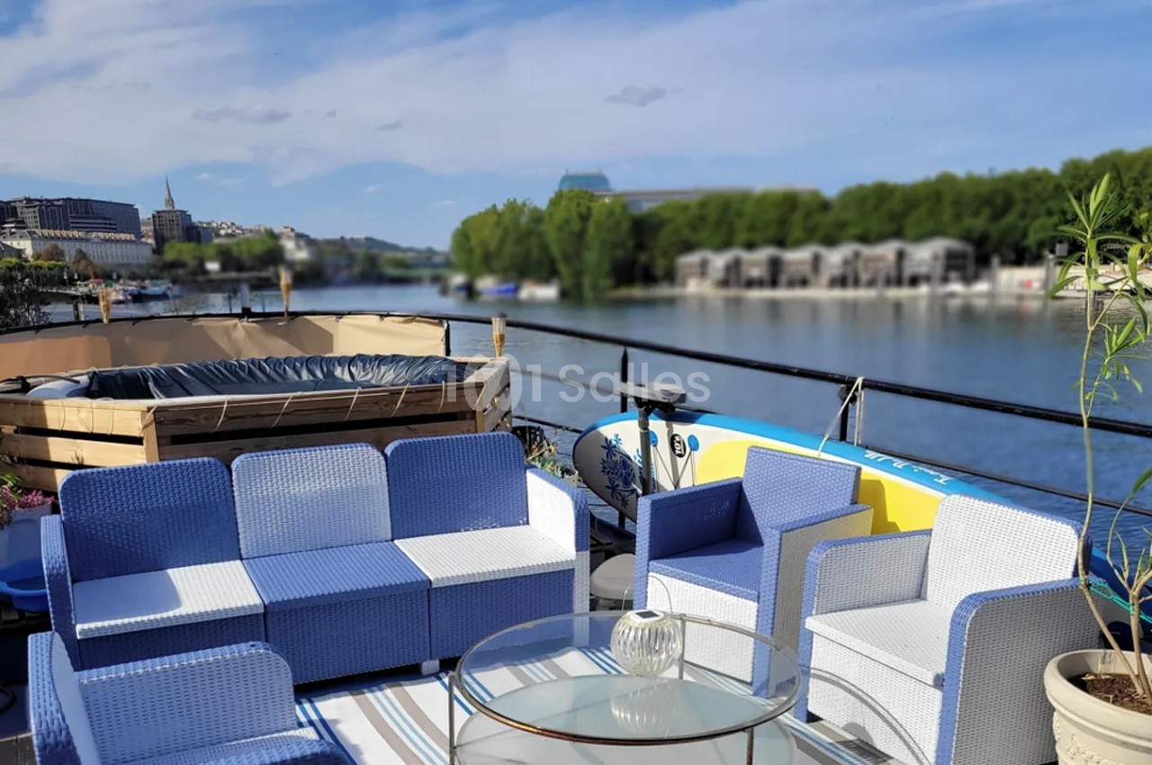 Mobilier de jardin bleu et blanc sur une terrasse au bord d'une rivière, avec jacuzzi et végétation en arrière-plan.