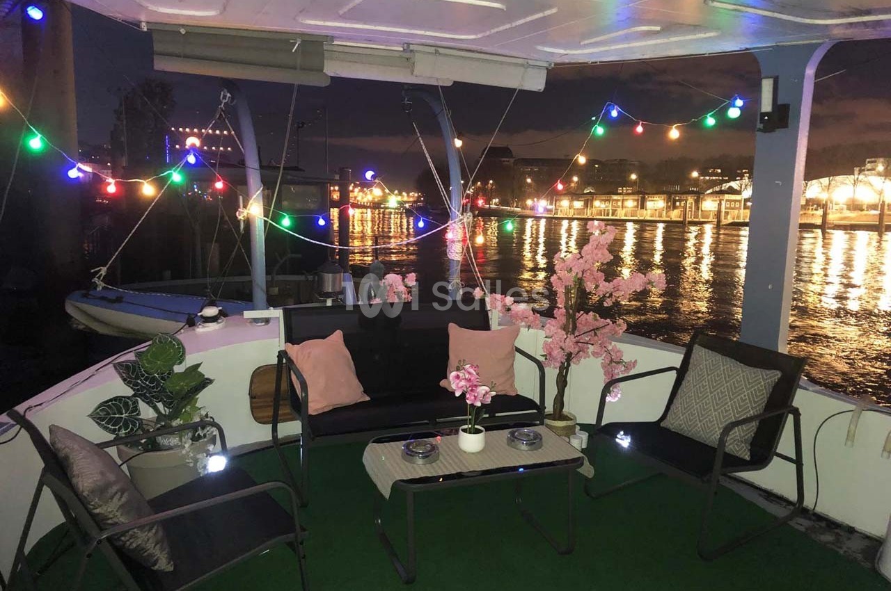 Espace extérieur d'un bateau aménagé avec des meubles, des plantes et des guirlandes lumineuses, vue sur un port éclairé.