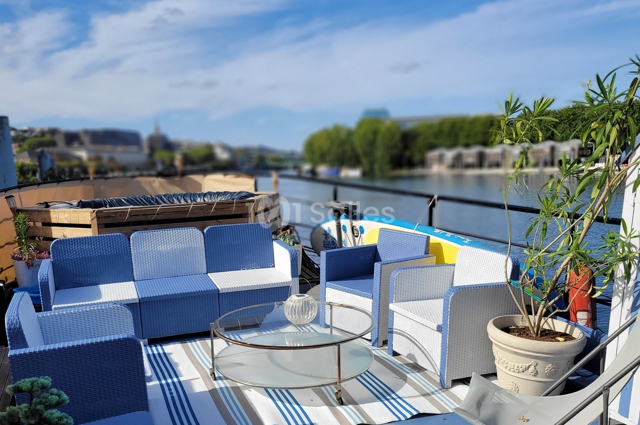 Salon de jardin avec canapés bleus et blancs sur une terrasse au bord de l'eau, entouré de plantes et d'une vue dégagée.