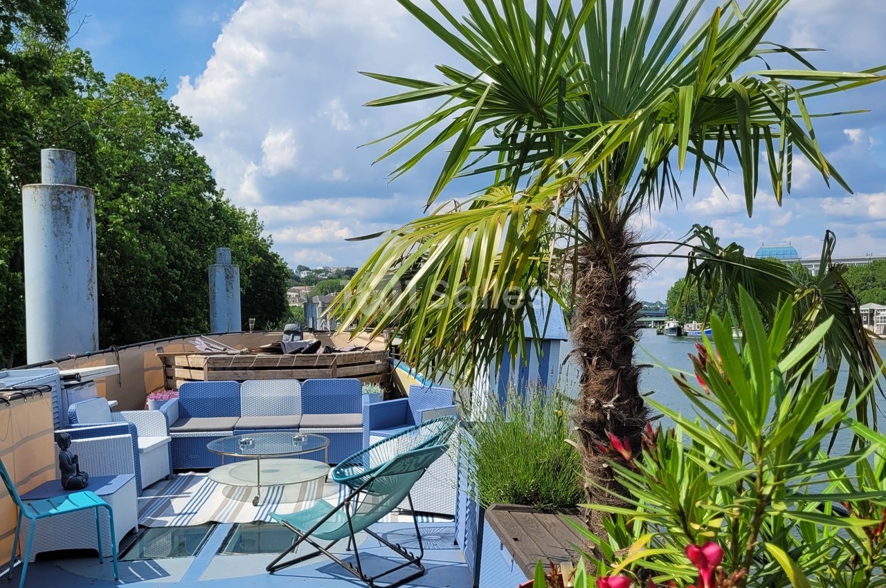Terrasse aménagée sur une péniche avec fauteuils, table basse, plantes et vue sur un fleuve bordé de verdure.