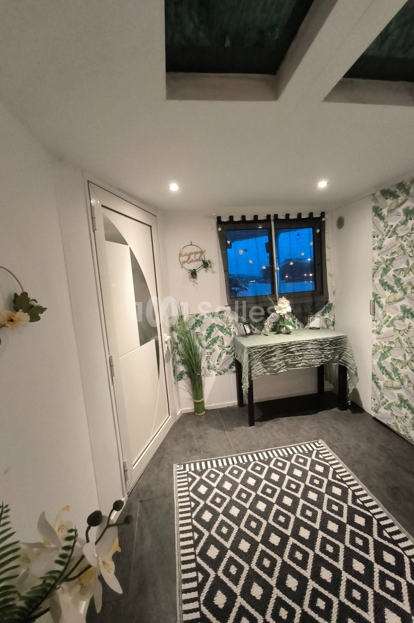 Intérieur lumineux avec décoration végétale, table ornée, tapis à motifs géométriques et fenêtre donnant sur l'extérieur.