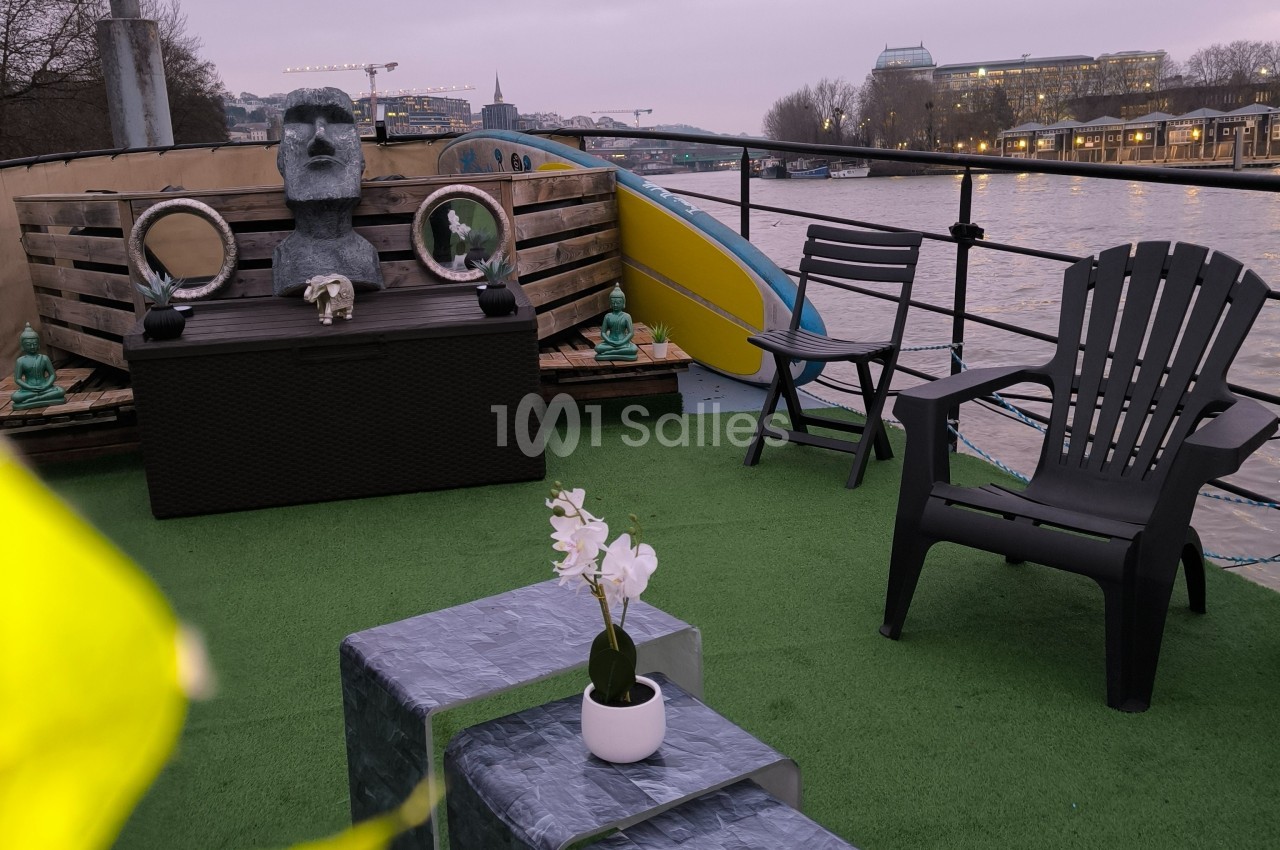 Terrasse aménagée sur une péniche avec fauteuils, table basse, orchidée, statue et vue sur un fleuve au crépuscule.