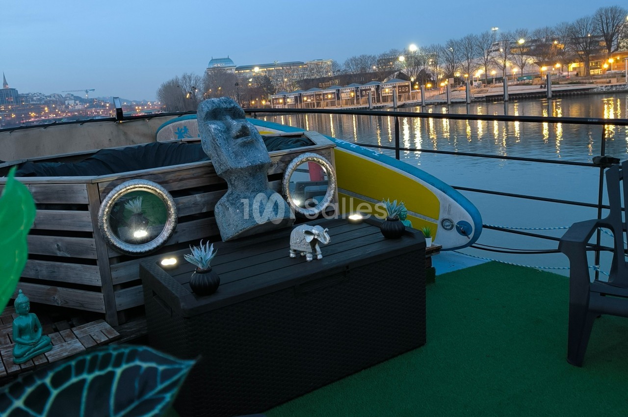 Statue et décorations sur une terrasse de péniche au bord d'un fleuve, avec des lumières et bâtiments en arrière-plan.