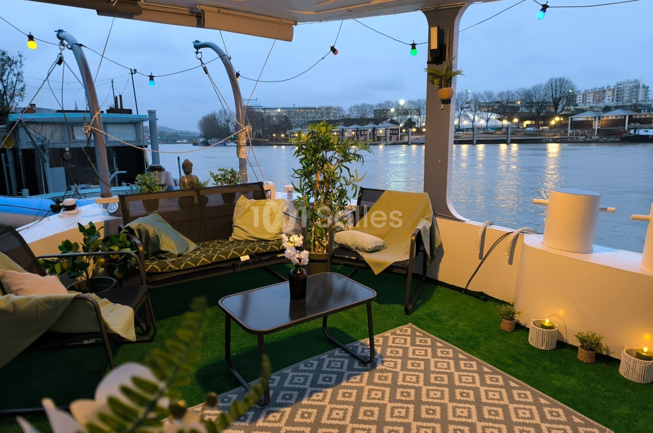 Terrasse aménagée sur une péniche avec fauteuils, table basse, plantes et vue sur une rivière au crépuscule.