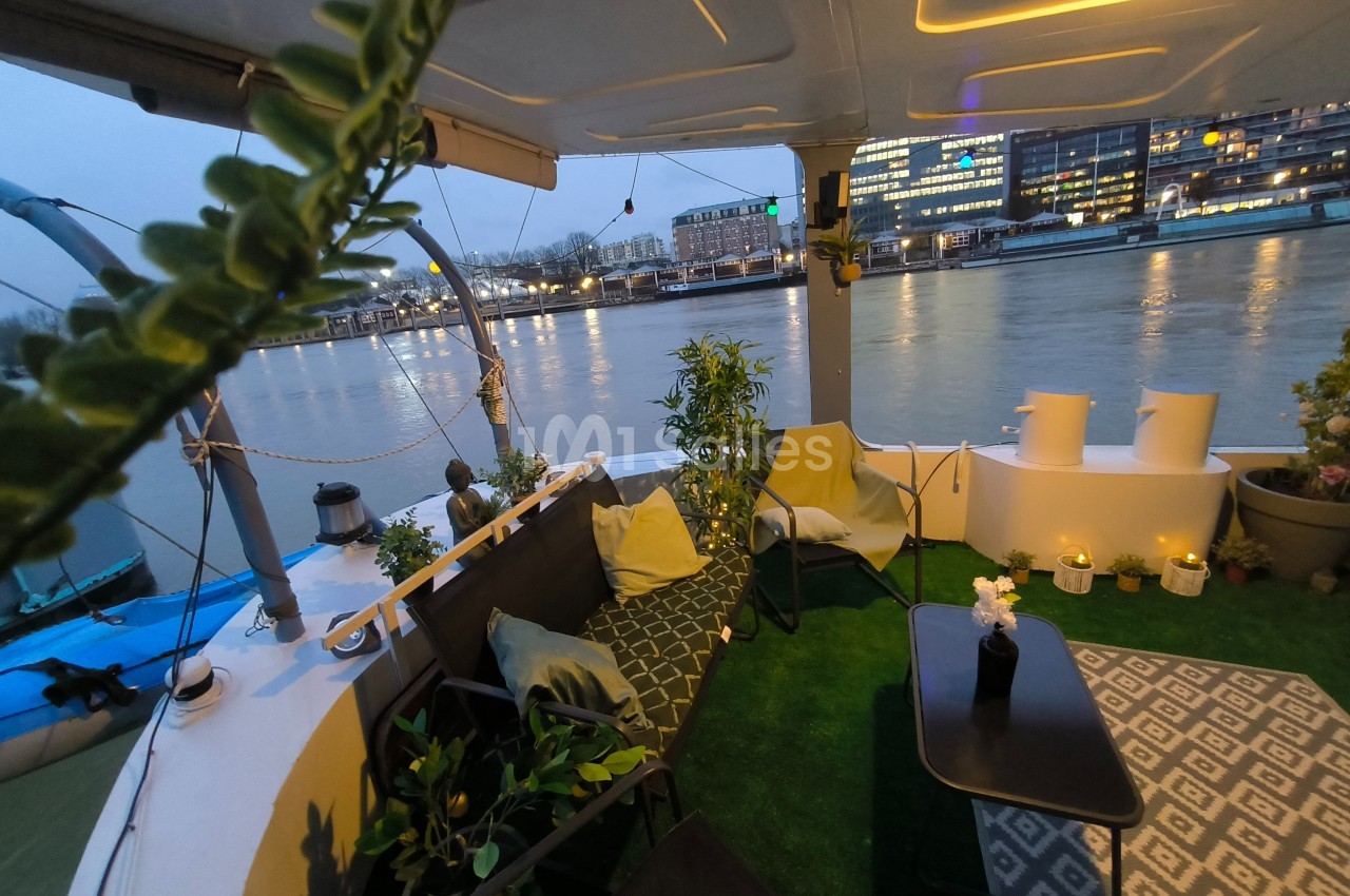 Terrasse aménagée sur un bateau avec vue sur une rivière et des bâtiments illuminés en arrière-plan.