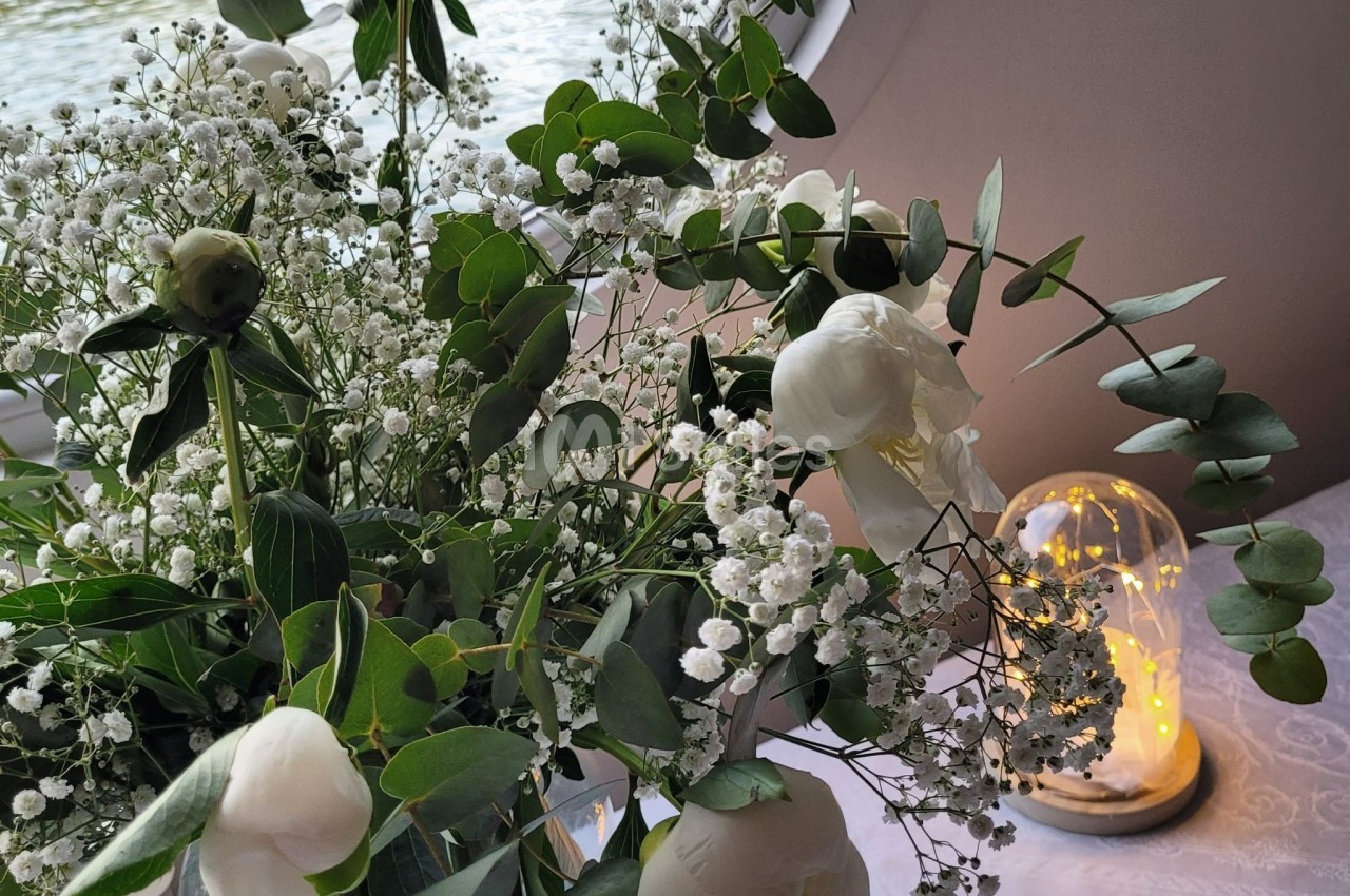 Bouquet de fleurs blanches et feuillage vert posé sur une table près d'une cloche lumineuse et d'une fenêtre.
