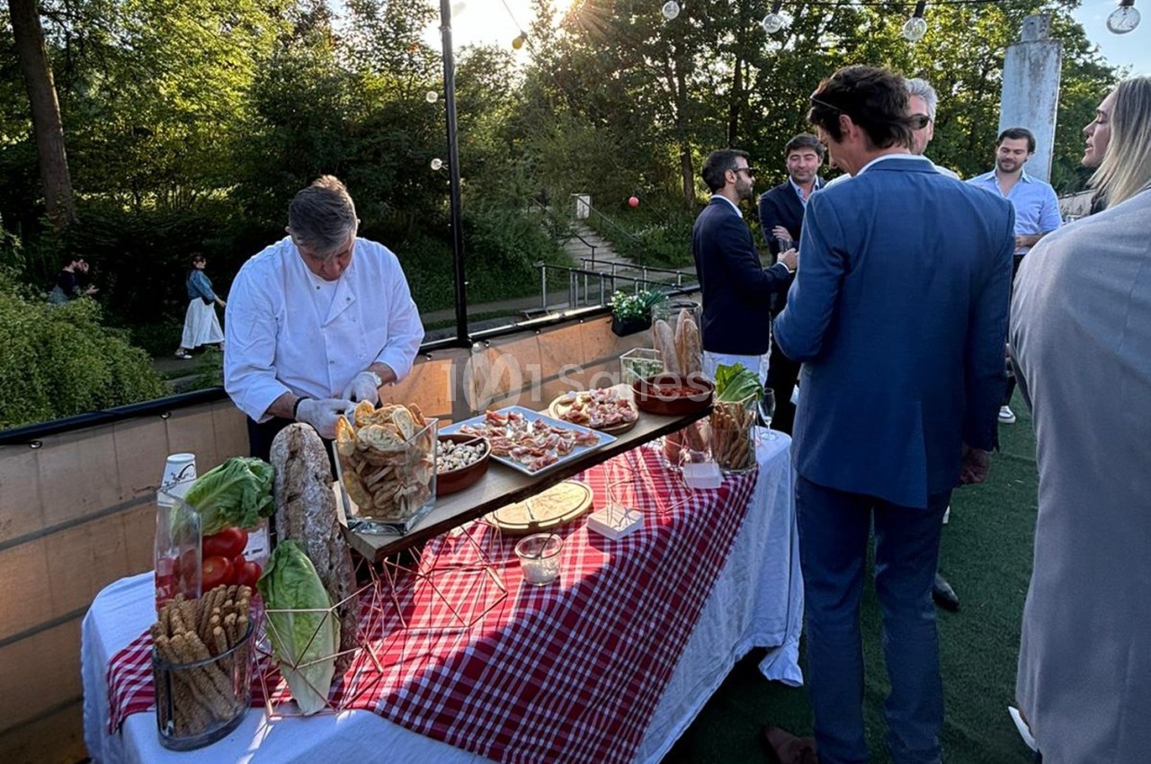 Un chef prépare des plats sur une table garnie de nourriture lors d'un événement en extérieur, entouré de convives.