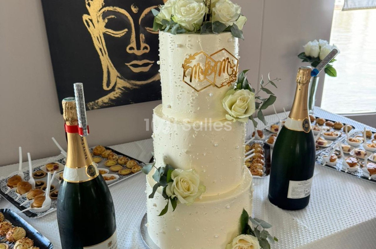 Gâteau de mariage à trois étages décoré de roses blanches, entouré de champagne et de desserts sur une table.