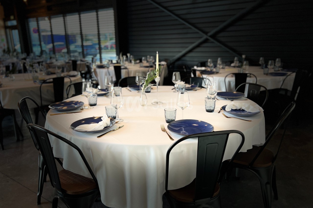 Salle de restaurant avec tables rondes dressées, nappes blanches, assiettes bleues et chaises noires.