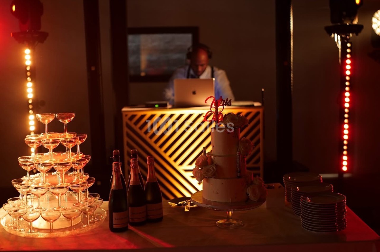 Un DJ mixe derrière une table éclairée, avec un gâteau, des coupes empilées et des bouteilles de champagne au premier plan.
