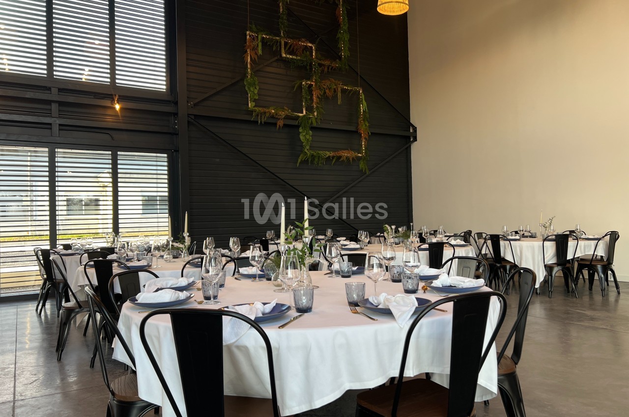 Salle de réception avec tables rondes dressées, nappes blanches, chaises noires et décoration murale végétale.